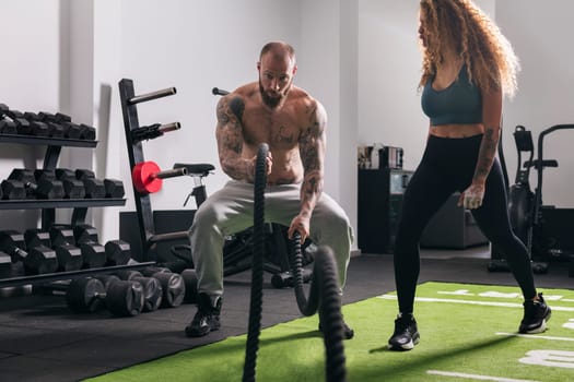 muscular sportsman with tattoos doing exercise with rope during functional workout with female instructor at gym, concept of fitness and active lifestyle