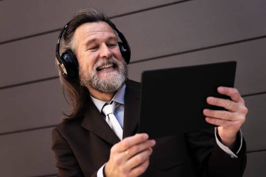 content mature male with beard in formal suit and headphones using tablet while laughing communicating online against gray wall, concept of middle age business man and technology of communication