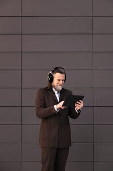 content mature male with beard in formal suit and wireless headphones using tablet while communicating online against gray wall, concept of middle age business man and technology of communication