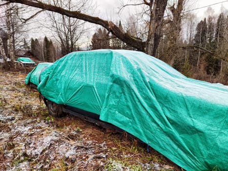 Tarps cover cars damaged in accidents, providing protection from weather elements in a wooded area during overcast conditions.