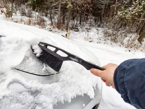 On a chilly winter day, a person removes snow from a car roof with a brush while surrounded by a snowy landscape.