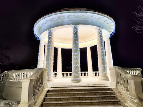 Illuminated gazebo with columns shines brightly in a snowy park during winter night, creating a serene and magical atmosphere.