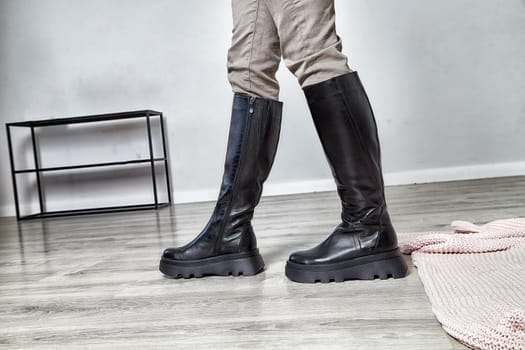 Model poses confidently in trendy black knee-high boots, set in a contemporary room with sleek decor.