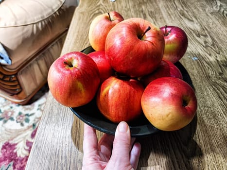 A hand holds a black bowl filled with vibrant red and yellow apples on a wooden table in a warm home setting.