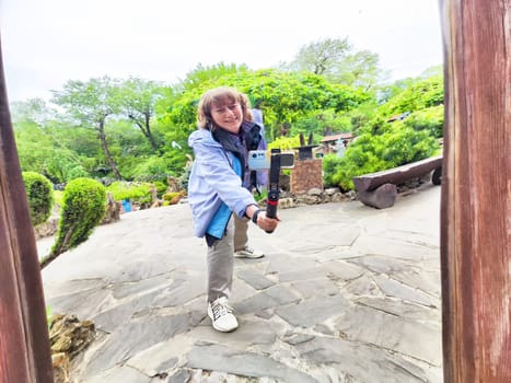 Middle-aged woman happily takes a selfie in a curved mirror while exploring a beautiful park filled with nature.