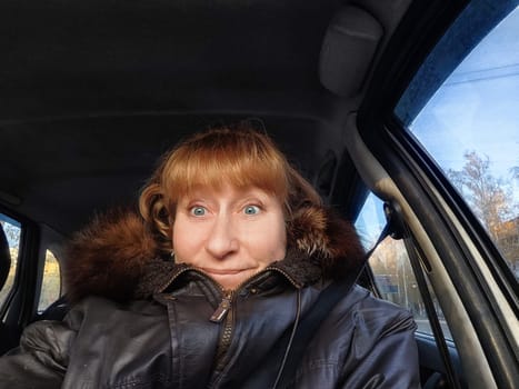A middle-aged woman smiles for a selfie in her car, capturing the joy of travel and adventure on a sunny morning journey.