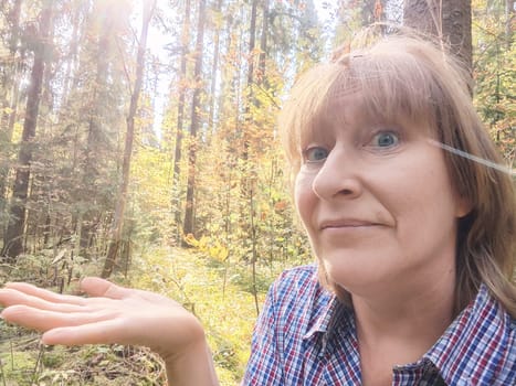 A middle-aged blonde woman is having fun in the forest, making funny faces as she explores the vibrant surroundings.