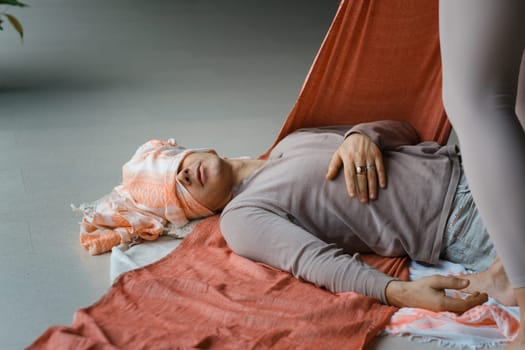 A peaceful indoor setting features a person lying on a mat, supported by a soft fabric. Another individual gently adjusts the fabric, aiding in relaxation while the atmosphere fosters a sense of calm and healing.