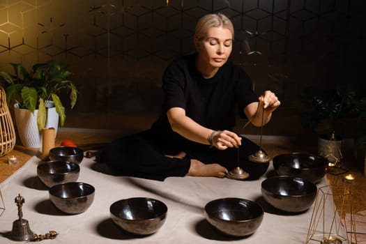 A woman sits cross-legged on a soft surface, carefully using a pendulum above several singing bowls. The calm indoor atmosphere features soft lighting and plants, creating a peaceful setting for sound healing.