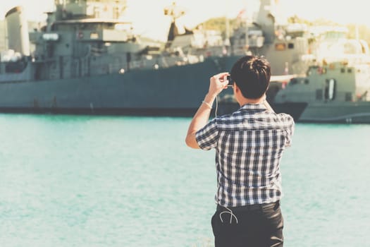 Asian man travel at navy harbor and take a photo warship or battleship by his smartphont at navy port with sea and blue sky view
