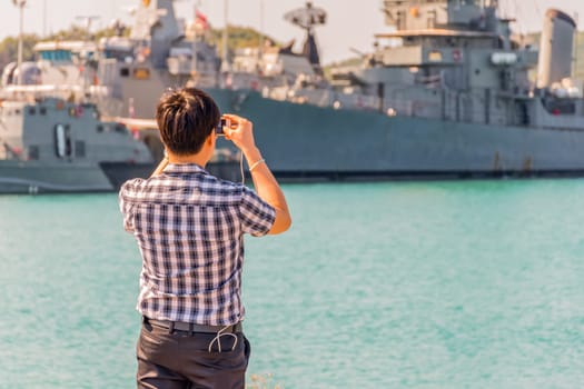Asian man travel at navy harbor and take a photo warship or battleship by his smartphont at navy port with sea and blue sky view