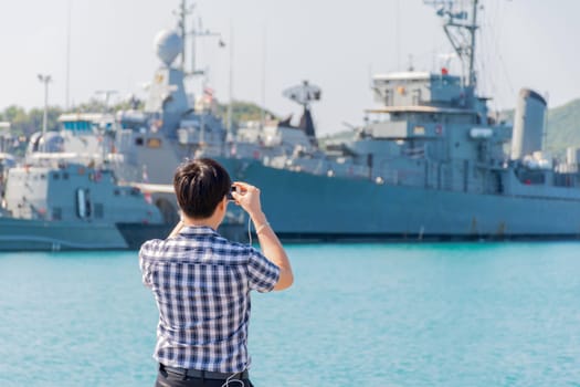 Asian man travel at navy harbor and take a photo warship or battleship by his smartphont at navy port with sea and blue sky view