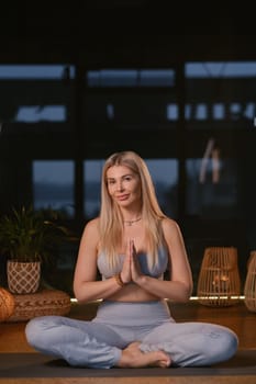 A woman is engaged in a yoga practice within a contemporary studio, surrounded by ambient lighting and decorative elements. She sits cross-legged on a mat, hands positioned in prayer, embodying tranquility and focus.
