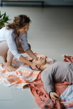 A man is lying on the floor, wearing casual clothes, as another person gently ties a cloth around his eyes. Soft fabrics are layered around him, creating a calming atmosphere perfect for relaxation or meditation.
