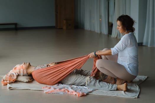 Two individuals engage in a therapeutic body treatment session in a calm studio. One person lies down, while the other gently pulls a soft fabric to aid in relaxation. The bright and serene surroundings enhance the soothing atmosphere.