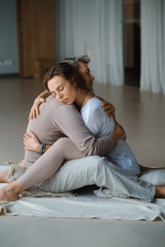 Two individuals sit closely on a soft mat, embracing each other in a heartfelt hug. The calm indoor setting features soft, natural light, creating a serene atmosphere conducive to emotional connection and wellness.