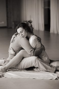 Two individuals sit closely on a soft mat, embracing each other in a heartfelt hug. The calm indoor setting features soft, natural light, creating a serene atmosphere conducive to emotional connection and wellness.
