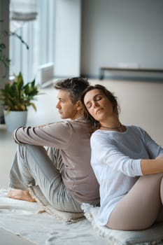 A man and woman sit back to back on a soft mat, surrounded by a tranquil indoor environment. The warm afternoon sunlight filters through large windows, creating a peaceful atmosphere as they focus on their mindfulness meditation practice together.
