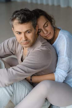A man and woman sit back to back on a soft mat, surrounded by a tranquil indoor environment. The warm afternoon sunlight filters through large windows, creating a peaceful atmosphere as they focus on their mindfulness meditation practice together.