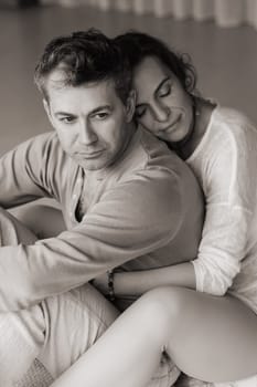 A man and woman sit back to back on a soft mat, surrounded by a tranquil indoor environment. The warm afternoon sunlight filters through large windows, creating a peaceful atmosphere as they focus on their mindfulness meditation practice together.