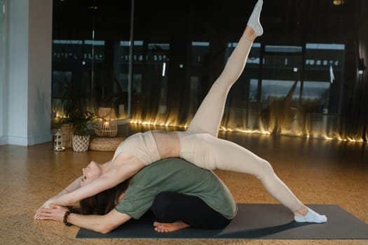 A woman balances gracefully in a difficult yoga pose while resting on the back of a man, who supports her with stability. The modern studio features large windows and soft lighting, creating a tranquil morning atmosphere ideal for yoga.