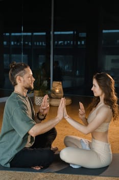 A couple sits cross-legged on mats in a serene yoga studio, gently pressing their palms together in a synchronized motion. The warm light highlights their calm expressions as they engage in a mindful practice, fostering a deep sense of connection.