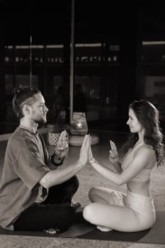 A couple sits cross-legged on mats in a serene yoga studio, gently pressing their palms together in a synchronized motion. The warm light highlights their calm expressions as they engage in a mindful practice, fostering a deep sense of connection.