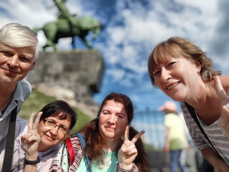 Four friends gather for a joyful selfie at a scenic outdoor location beneath a beautiful statue on a sunny day.