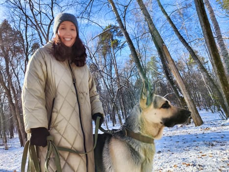 A middle-aged woman walks with her German Shepherd dog through a snowy park on a clear winter day, enjoying the fresh air and scenery.