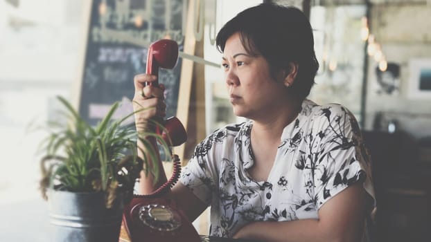 Asian women 40s white skin in black and white shirt unhappy and angry between waiting something or talking phone call in a coffee shop cafe with a red telephone vintage style