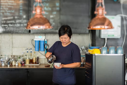 Asian woman 40s white skin work as a barista pouring latte froth to make a coffee latte art into the white coffee cup for serve to customers in the coffee shop cafe
