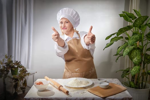 An elderly woman expertly prepares flour in a warm kitchen while playfully posing with a big smile and thumbs up.