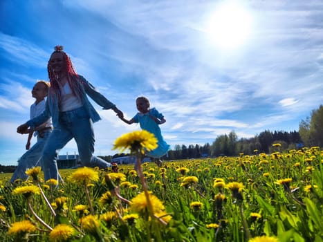 Children run happily alongside an adult in a sunny field filled with blooming dandelions under a clear blue sky.