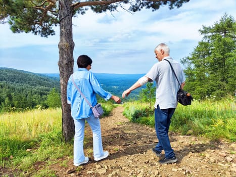 Two people stroll along a scenic trail, enjoying the view of lush hills and a blue sky on a sunny day.