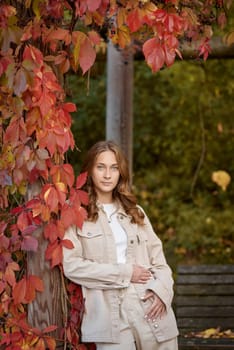 A young woman with wavy light brown hair and a cheerful smile is leaning against a wooden pillar wrapped in red autumn leaves. She is dressed in a beige casual outfit, including a stylish denim jacket and matching pants, with a white top underneath. The background features a scenic garden pergola with wooden beams covered in lush green and red foliage, creating a natural and cozy atmosphere. The warm lighting and soft depth of field enhance the autumn aesthetic, making this image perfect for seasonal fashion, lifestyle blogs, outdoor portraiture, and autumn-themed marketing campaigns.