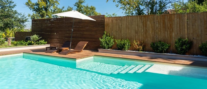 Beautiful backyard swimming pool with turquoise water, wooden deck, lounge chairs, umbrella, and greenery