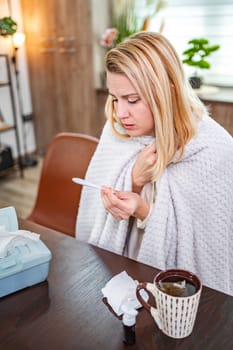 A woman checks her temperature at a table, wrapped in a blanket.