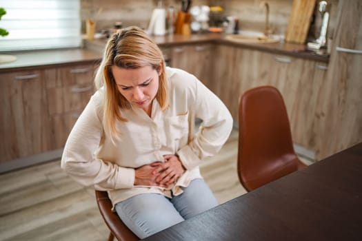 Woman sits in a rustic kitchen, holding her stomach in discomfort.