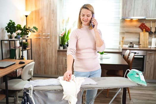 Woman balances phone call while ironing clothes in modern kitchen space