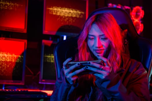 Gamer playing online game application on mobile phone wear gaming headphones, Asian woman live stream she play video game via smartphone at home neon lights living room