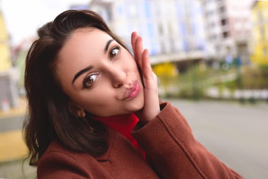 Cute young stylish woman in a brown autumn coat takes a selfie on the street for her boyfriend.