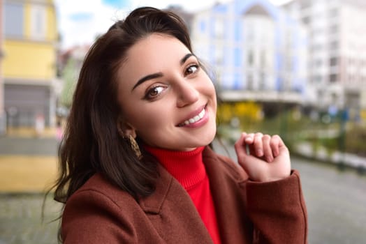 Portrait of beautiful lady in a brown autumn coat smiling while talking selfie on the street.