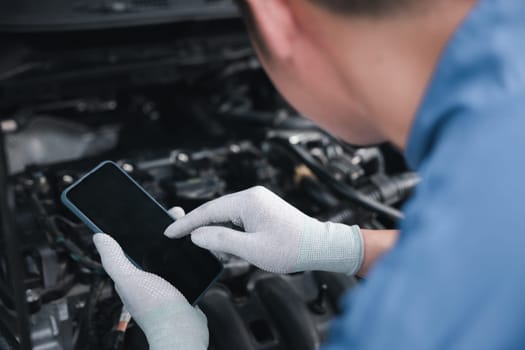 Male engineer using creativity and expertise to fix car engine at repair shop. Close-up shot of mechanic's hand holding wrench and mobile phone. Horizontal photo with garage background.