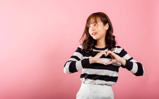 Portrait of a cheerful Asian woman making a heart symbol with two hands, expressing happiness on a pink background. Sending love and joy for Valentine's Day with confidence.