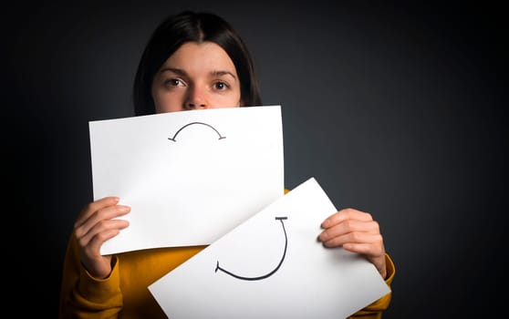 Girl changes, hides emotions, holds 2 papers with sadness and joy, the concept of duality and removal of masks, a woman removes her smiley mask and shows her difficulties with depression.