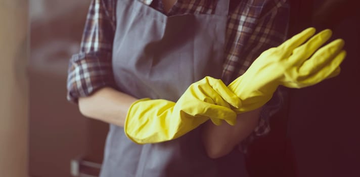 A young girl in a plaid shirt and apron puts yellow rubber gloves on her hands to start cleaning her house and create comfort. Housekeeper with gloves doing disinfection.