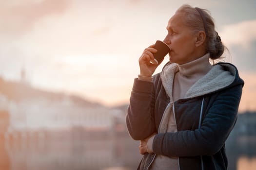 An adult beautiful woman holds a glass of aromatic coffee in her hands and enjoys the taste and atmosphere at sunset, views of the city and the river.