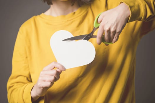 A girl holds scissors in her hands and cuts a heart, a young woman in a yellow sweater expresses her feelings, ends an abusive relationship with toxic love, dissolves a marriage, hands closeup.