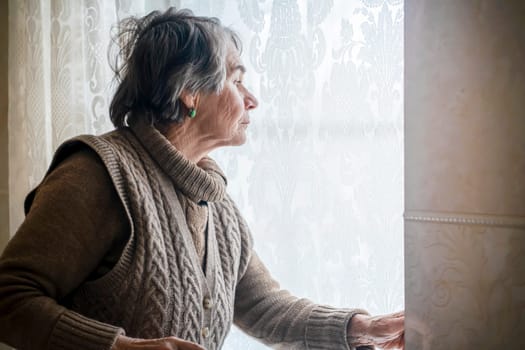 An elderly gray-haired woman in a casual knitted sweater looks out the window of her house, an old grandmother is waiting for guests, stands against the background of a window with lace curtains.