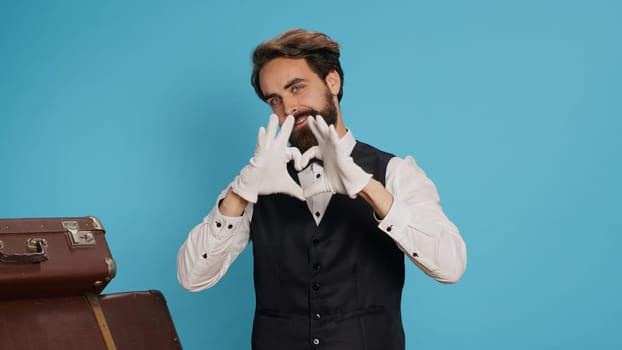 Cheerful bellhop does heart shaped sign in studio, expressing romantic honest feelings and flirting while he wears formal suit. Lovely employee showing romance symbol for love, sincere emotions.